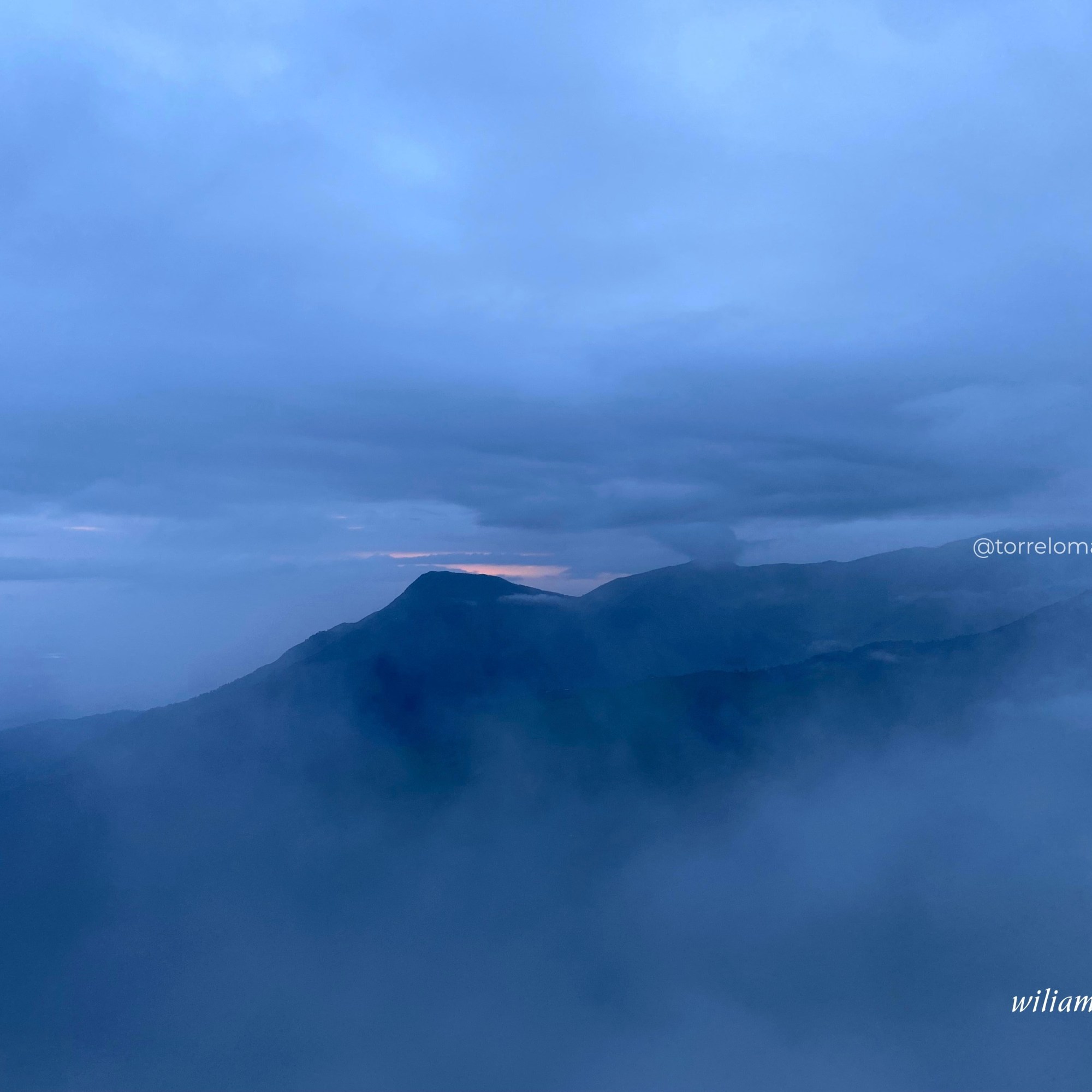 Silueta de una cordillera montañosa en tonos azules oscuros bajo un cielo nublado azul profundo, con un pequeño destello de luz naranja de un amanecer o atardecer en el horizonte.