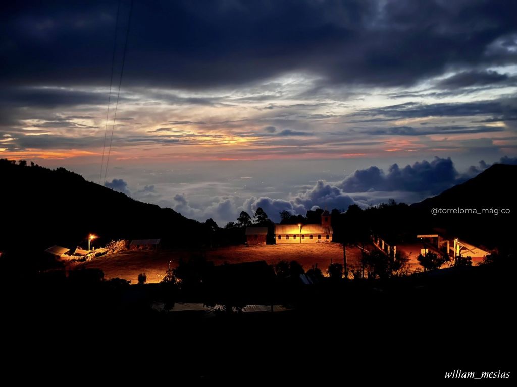 Vista nocturna de la comunidad de Torreloma iluminada entre montañas con un manto de nubes al fondo bajo un cielo crepuscular